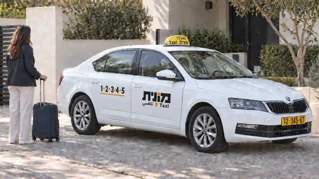 נוסעת עם מזוודה כחולה כהה לצד מונית ישראלית לבנה בכניסה לבית מגורים.