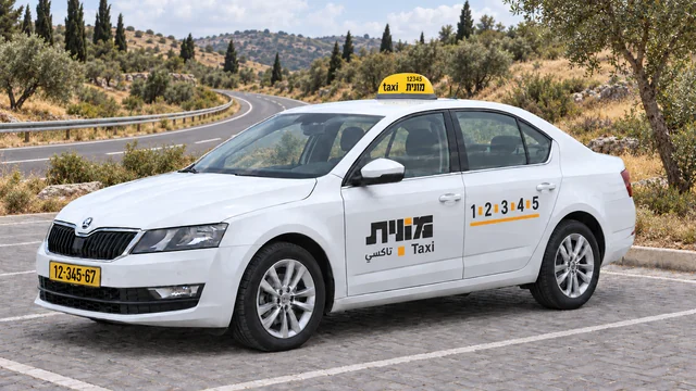 מונית ישראלית לבנה ברחבה מרוצפת ליד כביש מתעקל ונוף גבעות.