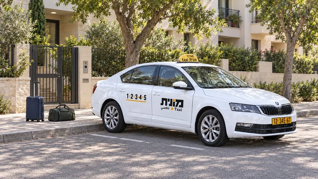 מונית ישראלית לבנה ליד שער מתכת וכניסה לבניין מגורים, עם מזוודה כחולה כהה ותיק נסיעות ירוק על המדרכה.