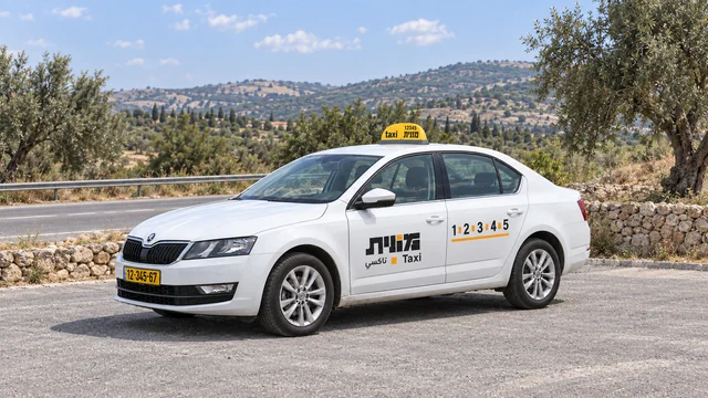 מונית ישראלית לבנה ברחבת עצירה ליד כביש, עצי זית, קיר אבן נמוך וגבעות ברקע.