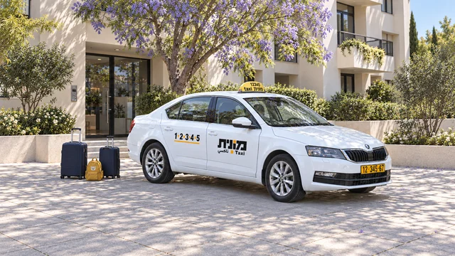 מונית לבנה לצד שתי מזוודות כחולות כהות ותיק גב צהוב ליד כניסה לבניין מגורים.
